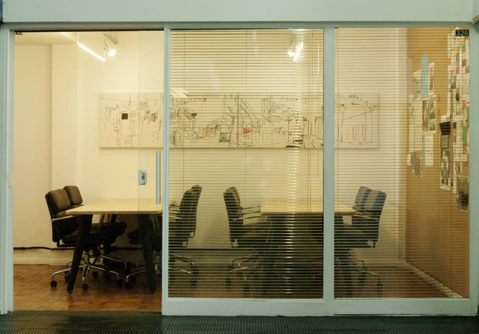 Foto do escritório do InternetLab, em que há uma porta de correr de vidro, duas mesas brancas com quatro cadeiras pretas em cada uma. Na parede, há uma pintura branca, preta e laranja do centro de São Paulo.