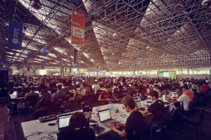 Image of a shed with many long, white, shared tables. At the tables, people are sitting using computers.
