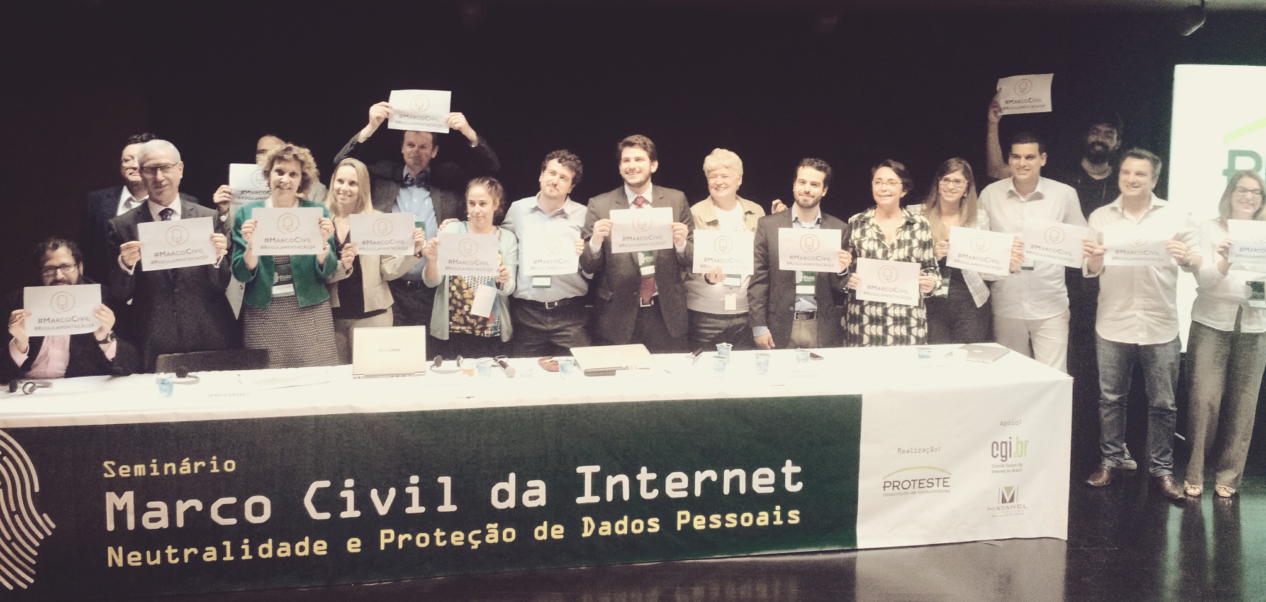 Foto de 18 pessoas em pé, segurando um papel, atrás de uma bancada em que está escrito: "Seminário: Marco Civil da Internet Neutralidade e Proteção de Dados pessoais". 