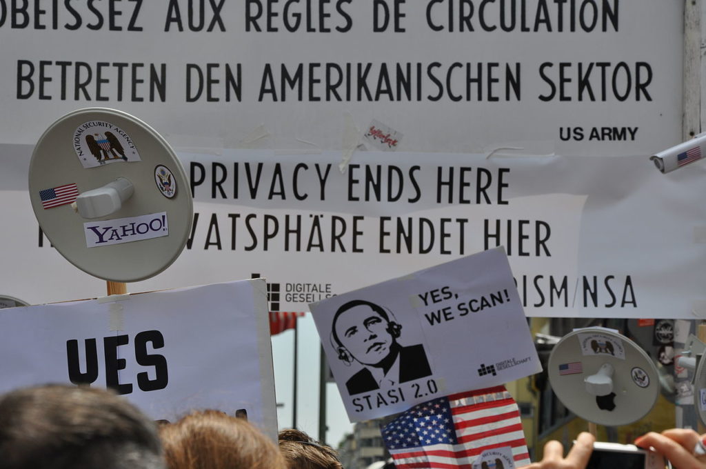 Photo of white posters with black letters, in a protest. The posters are in German. In the foreground, there is a small poster with an illustration of Barack Obama, wearing a headset, with the words: "yes, we scan!".